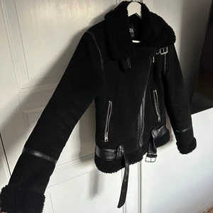 Svart jacka med pälsdetaljer Zara - Snygg svart jacka med pälsdetaljer och dragkedja. Jackan har ett bälte i midjan och långa ärmar. Perfekt för kyligare dagar med sin stilrena design. Från Zara