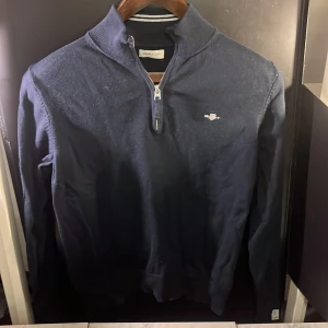 Mörklblå zip GANT tröja - Snygg mörkblå tröja från GANT med dragkedja och ribbad krage. Perfekt för kyligare dagar med sin stilrena design och diskreta logga på bröstet. Har vecklats ihop lite vid midjan annars fint skick. Hör av er vid minsta fundering🙌