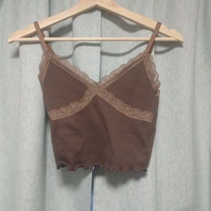 Brun ribbad linnetopp - Säljer en brun ribbad linnetopp från H&M med spetsdetaljer längs V ringningen. Toppen har justerbara axelband och en vågig kant nedtill. Perfekt för en stilren och bekväm look.