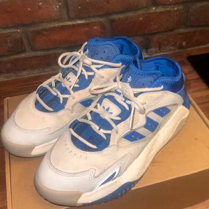 Blå och vita sneakers från Adidas - Snygga blå och vita unisex sneakers från Adidas med en unik design och snörning. Skorna har en robust sula och är perfekta för en sportig look. De har en kombination av blått och vitt med detaljer som ger dem en modern touch.