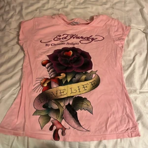 Rosa t-shirt från Ed Hardy - Säljer en rosa t-shirt från Ed Hardy med ett stort tryck av en ros och texten 'Love Life' på framsidan. T-shirten har korta ärmar och är gjord i bomull. OBS! Vet ej om den är äkta!