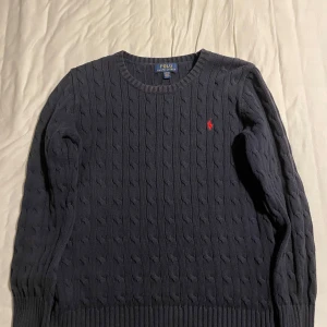 Mörkblå stickad tröja från Ralph Lauren - Säljer en stilren mörkblå stickad tröja från Ralph Lauren med kabelstickat mönster. Tröjan har långa ärmar och en liten röd logotyp på bröstet. Perfekt för kyliga dagar. Storlek L för barn, passar som storlek S (herr). Skriv gärna om ni har frågor angående vad som 