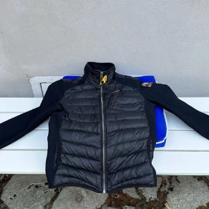 Svart pufferjacka från Parajumpers - Snygg svart cardigan från Parajumpers med dragkedja och hög krage. Jackan har långa ärmar och en cool patch på axeln. Perfekt för kyligare dagar. Skick 10/10 knappast använd!