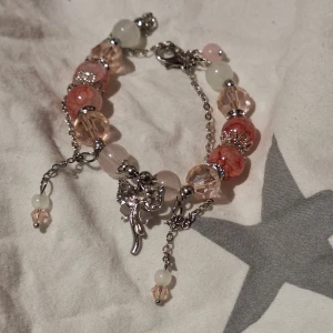 Charmigt armband med pärlor och berlocker - Säljer ett charmigt armband med pärlor i rosa och vita nyanser. Armbandet har flera detaljerade berlocker och en justerbar kedja i silverfärg. Perfekt för att ge en extra touch till din outfit.