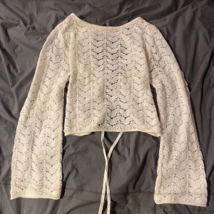 Vit stickad tröja med snörning - Säljer en vit stickad tröja med ett snyggt mönster och öppen rygg med snörning. Tröjan har långa ärmar och en croppad passform. Perfekt för en stilren look! Ärmarna är lite utsvängda, sitter jättesnyggt på! 