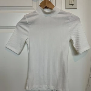 Vit ribbad polotopp från Kappahl - Säljer en stilren vit ribbad polotopp från Kappahl i storlek XS. Helt oanvänd. Toppen har korta ärmar och en hög krage, perfekt för en minimalistisk look.
