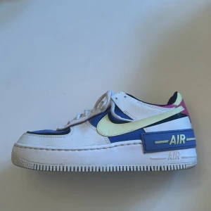 Nike Air Force med färgglada detaljer - Snygga Nike Air Force sneakers med en mix av färger som vit, blå, gul och rosa. Skorna har en klassisk snörning och en robust sula med 'AIR' märkning på sidan. Perfekta för en färgglad och sportig stil.