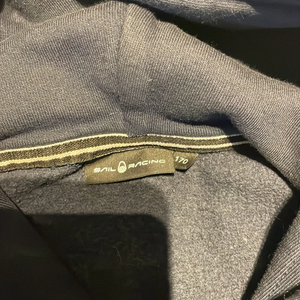 Säljer en mörkblå hoodie från Sail Racing med vit logga på bröstet. Tröjan har en klassisk design med huva och långa ärmar. Perfekt för en avslappnad stil.. Neuletakit & Villapaidat.