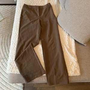 Bruna kostymbyxor från ONLY - Snygga bruna kostymbyxor från ONLY med en klassisk design. Byxorna har en rak passform och stängs med knapp och dragkedja. Perfekta för en stilren look.