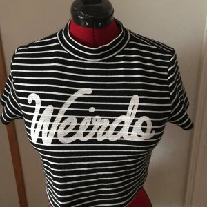 Randig topp med text från H&M - Säljer en svart och vit randig topp från H&M med texten 'Weirdo' i vitt på framsidan. Toppen har korta ärmar och en hög krage, perfekt för en avslappnad stil.