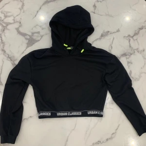 Svart croppad hoodie från Urban Classics - Säljer en svart croppad hoodie från Urban Classics med långa ärmar och huva. Den har en elastisk kant med Urban Classics-logga och neongröna detaljer vid dragsnörena.