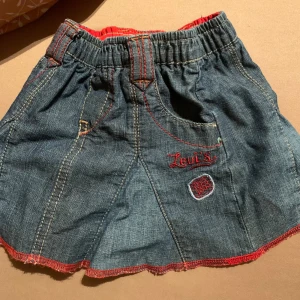 Jeanskjol från Levi's - Söt jeanskjol från Levi's med röda sömmar och broderad logga. Kjolen har elastisk midja och dekorativa fickor med nitar. Perfekt för små fashionistas!