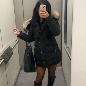 Svart dunjacka med päls från Hollies - Snygg svart dunjacka från Hollies med avtagbar päls på huvan. Jackan har dragkedja och knappar framtill samt praktiska fickor med dragkedjor. Väldigt varm. Nypris: 5300. Strl 36