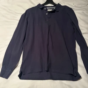 Mörkblå pikétröja från Pier One - Säljer en stilren mörkblå pikétröja från Pier One. Tröjan har långa ärmar och en klassisk krage med knappar framtill. Perfekt för en avslappnad men ändå snygg look.