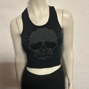 Svart croppad linnetopp med dödskallemotiv - Snygg svart croppad linnetopp med ett coolt dödskallemotiv i glittrande stenar på framsidan. Perfekt för en edgy look.