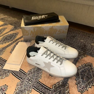 Vita sneakers från Golden Goose - Snygga vita sneakers från Golden Goose med grå stjärndetalj på sidan och svarta hälpartier. Skorna har grå snörning och en klassisk design som passar perfekt för en stilren look.