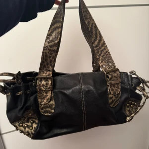 Svart axelväska med leoparddetaljer - Snygg svart axelväska med leopardmönstrade handtag och detaljer. Väskan har dragkedja och nitar på sidorna som ger en cool look. Perfekt för att ge din outfit en extra edge. Priset går att diskutera vid snabb affär