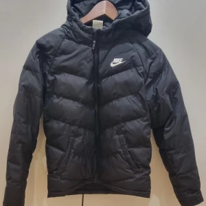 Svart dunjacka från Nike - Säljer en svart dunjacka från Nike med huva och dragkedja. Jackan har en klassisk pufferdesign och Nike-logga på bröstet. Perfekt för kyliga dagar. Junior stl L 147-158