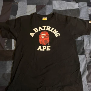 Svart t-shirt från A Bathing Ape - Säljer en svart t-shirt från A Bathing Ape med det ikoniska röda kamouflagemönstret och logotypen på bröstet. T-shirten har korta ärmar och en rund halsringning. Perfekt för streetwear-entusiaster!