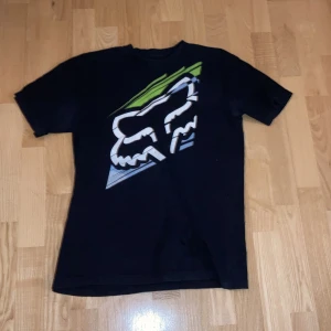 Svart fox tröja  - Snygg svart t-shirt från Fox med ett stort grafiskt tryck i vitt, grönt och blått på framsidan. Tillverkad i 100% bomull för en skön känsla. Perfekt för en avslappnad stil.