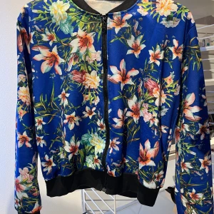 Blommig tunn bomberjackastil kornblå - Snygg blå bomberjacka med färgglatt blommönster. Jackan har svarta muddar och dragkedja framtill. Jackan hae 2 fickor.  Perfekt för att liva upp garderoben med lite färg och stil.