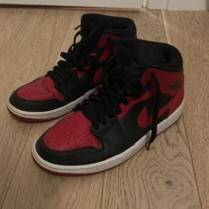 Nike Air Jordans 1 Mid  - Snygga Nike Air Jordans i klassisk röd och svart färgkombination. Skorna har hög snörning och en ikonisk design med Nike-loggan på sidan. Perfekta för sneaker-entusiaster.