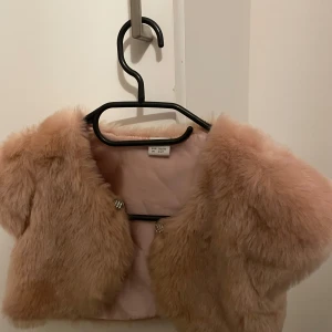 Rosa pälsjacka från Lindex Kids - Söt rosa pälsjacka från Lindex Kids, perfekt för att hålla sig varm och stilren. Jackan har en kort design med glittriga knappar framtill. Passar barn i åldern 4-6 år.