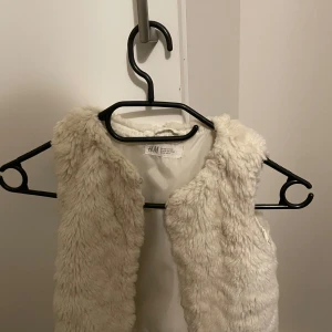 Vit pälsväst från H&M - Säljer en fluffig och mysig vit pälsväst från H&M. Perfekt för att hålla sig varm och stilren under kyligare dagar. Västen är ärmlös och har en mjuk känsla.
