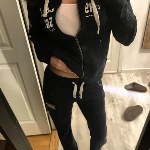 Mörkblå hoodie med dragkedja från Hollister - Säljer en mörkblå hoodie från Hollister med dragkedja och vit text på bröstet. Tröjan har en justerbar huva med vita snören och en amerikansk flagga på ärmen. Perfekt för en avslappnad stil.