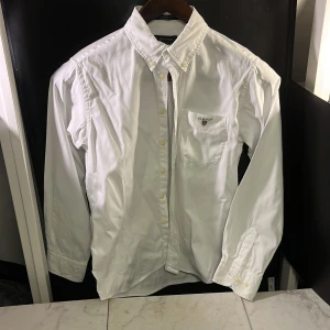 Vit skjorta från GANT - Säljer en klassisk vit skjorta från GANT i storlek 146-152. Skjortan har en bröstficka med broderad logotyp och knappar framtill. Perfekt för en stilren look, hör av er vid minsta fundering🙌