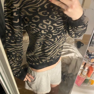 Svart och guldmönstrad tröja från ONLY - Säljer en snygg svart tröja från ONLY med ett djärvt leopardmönster i guld. Tröjan har långa ärmar och ribbade muddar vid ärmslut och nederkant. Perfekt för att ge din outfit en trendig touch! Tröjan är helt ny och inte andvänd. Det är storlek M men passar som S.