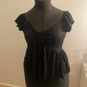 Svart topp med knyt och volang - Snygg svart topp med knyt framtill och volangdetaljer. Toppen har korta ärmar och en smickrande passform. Perfekt för en stilren look.