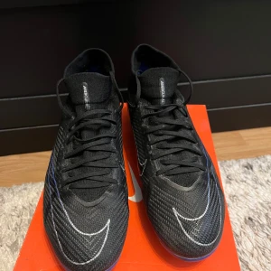 Nike Zoom Mercurial Superfly Pro FG - Snygga svarta Nike fotbollsskor med blå detaljer och snörning. Skorna har en modern design med en slitstark sula och är perfekta för fotbollsplanen. Har använt 5 gånger max på fotbollsplanen och känns jättesköna och foten har ett bra utrymme i skorna. size 44. Ny pris 1500kr. Pris kan diskuteras!