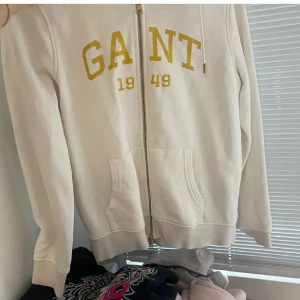 Vit hoodie från GANT - Säljer en stilren vit hoodie från GANT med dragkedja och gul text 'GANT 1949' på bröstet. Tröjan har långa ärmar och praktiska fickor framtill. Perfekt för en avslappnad look.