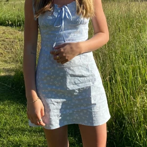 Ljusblå blommig klänning - Charmig ljusblå klänning med blommigt mönster och knytning framtill. Perfekt för sommarens varma dagar med sin korta längd och ärmlösa design.