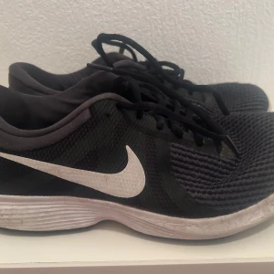 Svarta Nike sneakers - Säljer ett par svarta Nike sneakers med vit sula och vit swoosh-logga. Skorna har snörning och är perfekta för vardagsbruk.