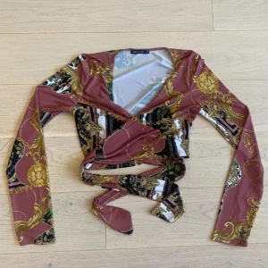 Mönstrad omlottopp från Nelly - Snygg mönstrad omlottopp från Nelly  i en mix av färger som rosa, guld och svart. Toppen har långa ärmar och ett knytband i midjan för en justerbar passform. Perfekt för att ge en trendig touch till din outfit. Använd 1 gång.