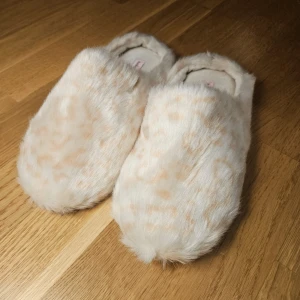 Beige fluffiga tofflor från Hunkemöller - Mysiga och fluffiga tofflor i beige med ett diskret leopardmönster från Hunkemöller. Perfekta för att hålla fötterna varma och bekväma hemma. Tofflorna har en mjuk insida och halkfri sula.