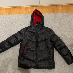 Svart pufferjacka från Closure LDN - Snygg svart pufferjacka från Closure LDN med röd insida och huva. Jackan har dragkedja och praktiska fickor med röda detaljer. Perfekt för kyliga dagar.