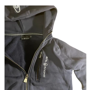 Mörkblå zip up från Sail Racing - Snygg blå jacka från Sail Racing med huva och dragkedja. Jackan har en bröstficka med dragkedja och mjukt foder. Perfekt för kyliga dagar. Däremot finns det små tydliga färg fläckar