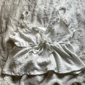 Vit ärmlös topp med volangdetaljer - Söt vit topp med volangdetaljer och knytband framtill. Perfekt för varma sommardagar. Den har en smickrande v-ringning och är tillverkad i ett lätt och luftigt material. Aldrig använd🩷