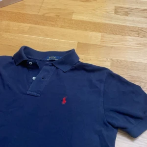  Ralph Lauren tröja - Säljer en klassisk mörkblå pikétröja från Polo Ralph Lauren med den ikoniska röda loggan broderad på bröstet. Tröjan har korta ärmar och en traditionell krage med knappar. Perfekt för en stilren och avslappnad look.