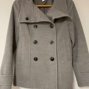 Grå kappa med dubbelknäppning - Säljer en stilren grå kappa i storlek 36 med dubbelknäppning framtill. Den har en klassisk design med två rader av knappar och en snygg detalj baktill med två knappar. Är dock isönder vid snöret som man kan hänga upp kappan med, utöver det finns det inga andra defekter. Perfekt jacka för höst och vår!