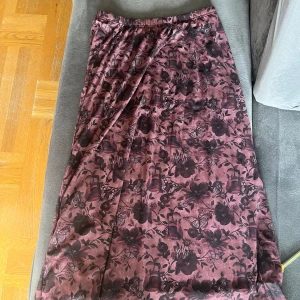 Blommig långkjol från Romwe - Säljer en superfin långkjol från Romwe med ett vackert blommigt mönster i svart och lila. Kjolen har en mjuk och flödande passform, perfekt för både vardag och fest. Den är gjord i ett lätt och bekvämt material som känns skönt mot huden. Kjolen är i nyskick och har aldrig använts, så den är redo för sin nya ägare!