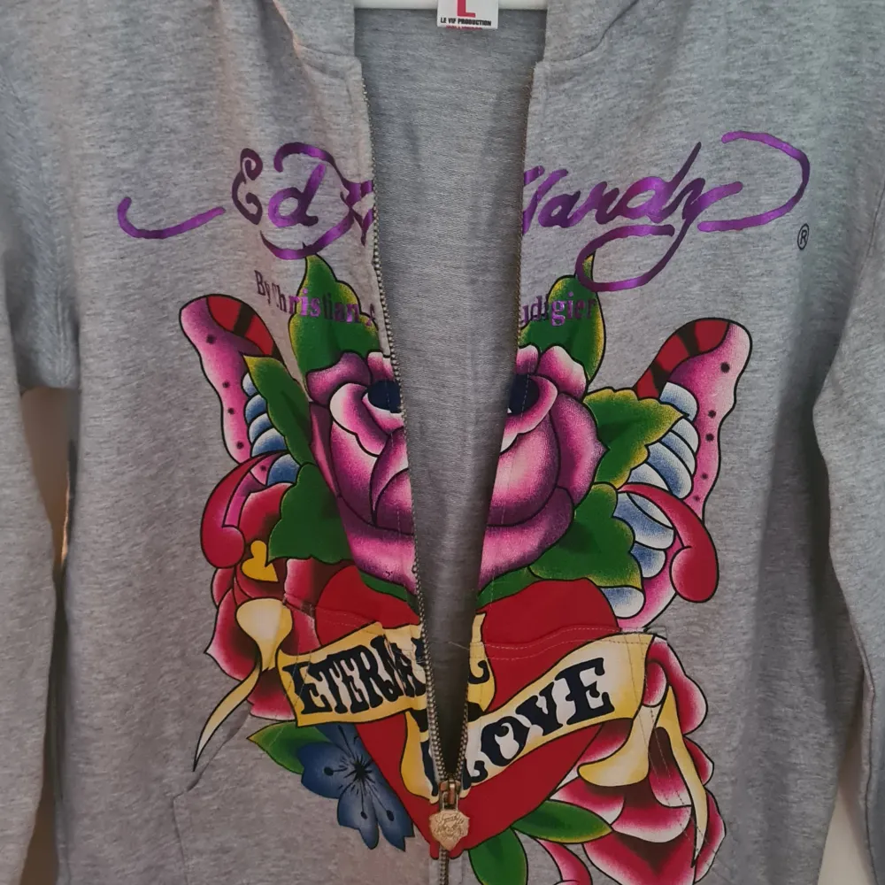 Säljer en grå hoodie från Ed Hardy med färgglada tatuering-inspirerade tryck. Den har en stor rosa ros och ett hjärta med texten 'Eternal Love' på framsidan. Hoodien har dragkedja och långa ärmar med tryck på båda ärmarna. 