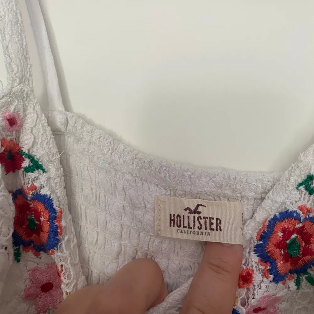 Säljer en superfin vit spetsklänning från Hollister. Den har ett vackert broderat blommönster i färgerna röd, blå och grön vid halsen. Klänningen är ärmlös och har en tight passform med band i midjan. 🌸. Klänningar.