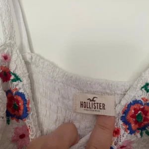 Vit spetsklänning från Hollister - Säljer en superfin vit spetsklänning från Hollister. Den har ett vackert broderat blommönster i färgerna röd, blå och grön vid halsen. Klänningen är ärmlös och har en tight passform med band i midjan. 🌸