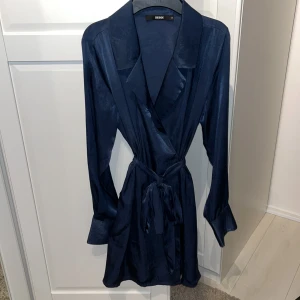 Mörkblå omlottklänning från Bik Bok - Säljer en elegant mörkblå omlottklänning från Bik Bok. Klänningen har långa ärmar och ett knytband i midjan för en smickrande passform. Materialet är glansigt och ger en lyxig känsla. Perfekt för festliga tillfällen!