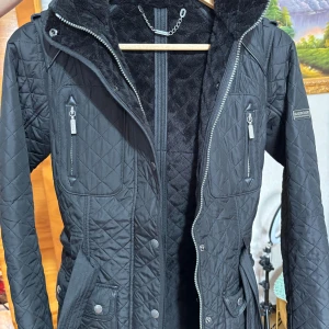 Svart quiltad jacka från Barbour - Säljer en snygg svart quiltad jacka från Barbour International. Jackan har en stilren design med dragkedja och knappar framtill. Den är långärmad och har flera praktiska fickor med dragkedjor. Perfekt för höst och vår!