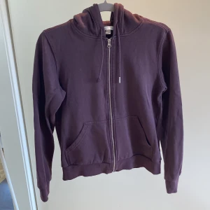 Vinröd zip-up från H&M - Säljer en mysig vinröd hoodie från H&M i storlek XS. Den har en dragkedja framtill och en praktisk huva med snören. Perfekt för kyliga dagar eller som ett extra lager. Den är i bra skick men den saknar ett metallhökje för ett av snörena. 